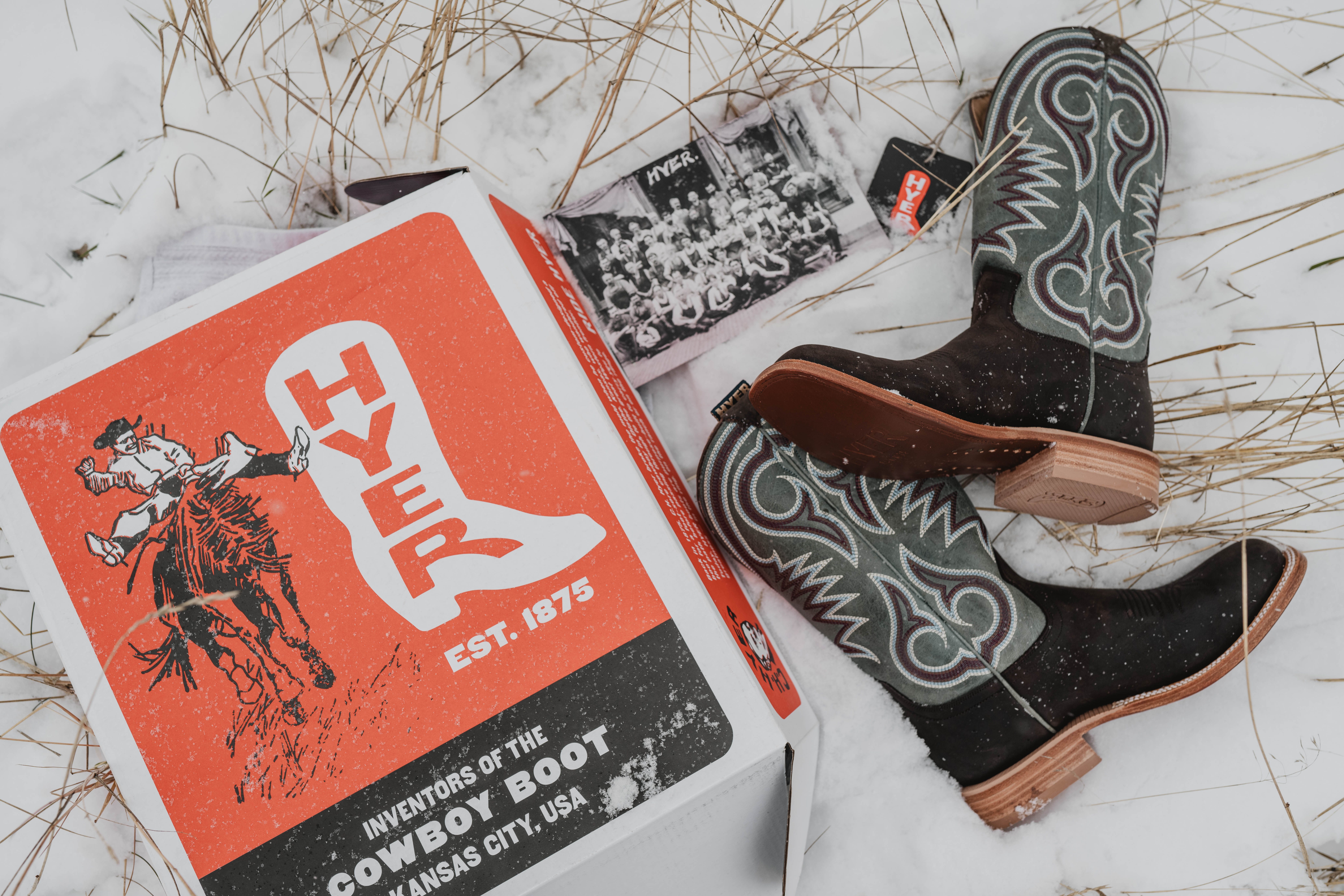 A winter layout of Hyer cowboy boots, their box and info card over snow and straw