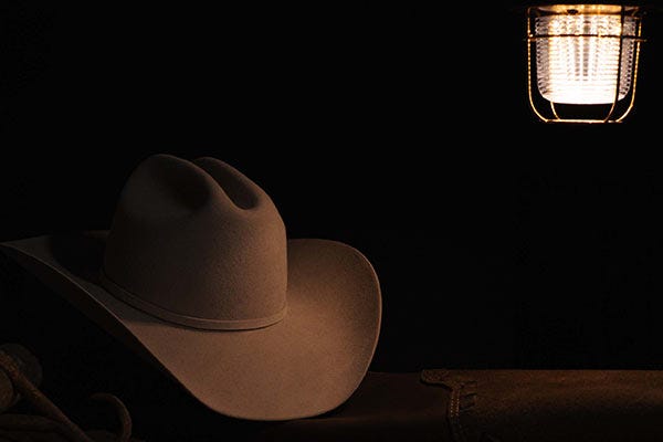 A straw cowboy hat sits in low light beneath a warm hanging lantern, creating a moody Western atmosphere against a black background.