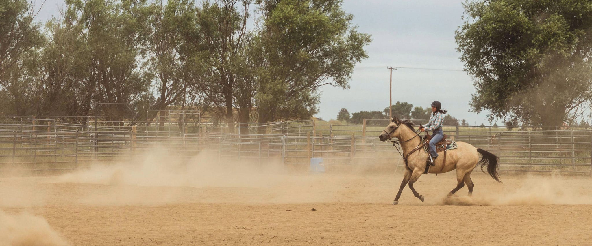 Girl on horse in dusty warm up area in jeans, helmet, a western top, boots and a helmet
