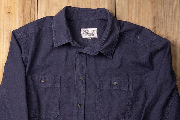 Dark blue Benton button-up shirt made from hemp fabric is displayed flat against a wooden background, featuring two front chest pockets, button closures, and a sewn collar.