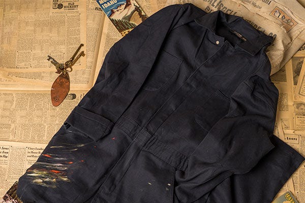 Dark blue work coveralls with paint marks on one leg, laid on a background of scattered vintage book pages and magazines.