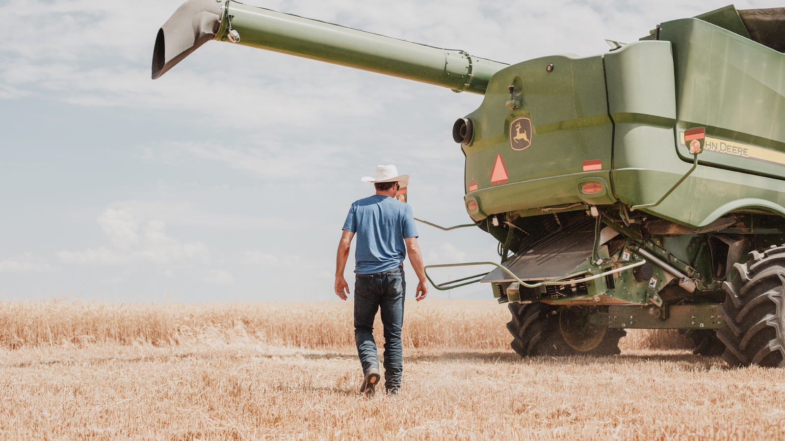 A man in a cowboy hat walking toward a John Deere combine combine in a wheat field