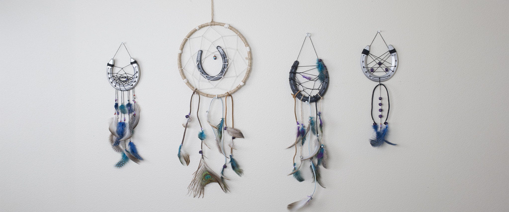 Three handmade dreamcatchers hanging on a wall, each featuring a metal horseshoe woven into the center and decorated with string and hanging feathers.