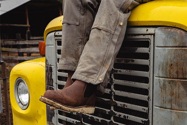 Brown work boots and worn gray work pants resting on the front grille of a yellow, weathered truck.