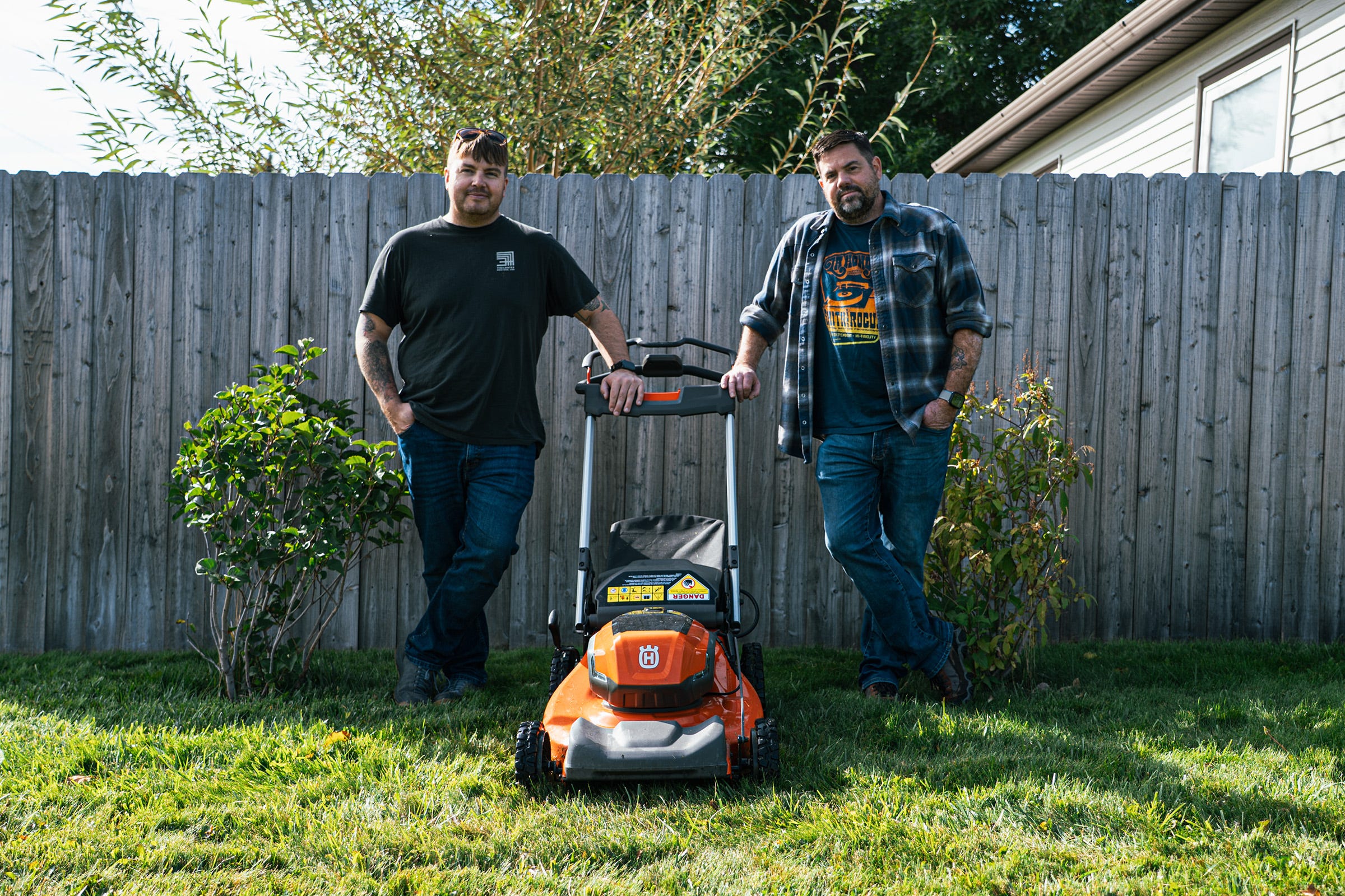 Husqvarna LE-322R battery-powered lawn mower positioned in a residential backyard during a hands-on product review, with two presenters standing behind the mower in front of a wooden privacy fence.