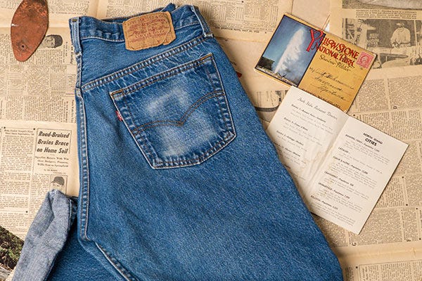Blue denim jeans with a back pocket and Levi leather patch, laid on a surface of vintage newspaper pages alongside postcards and printed booklets.