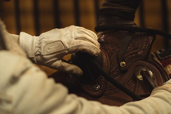 A man loads a saddle on a horse while wearing Noble Outfitters leather work gloves.