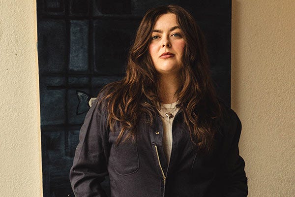 Maddie wearing a Berne coveralls over a light waffle shirt, standing indoors in front of a textured wall and a dark window.