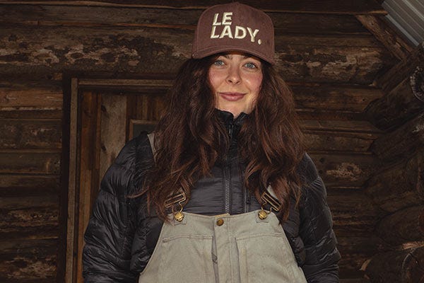 Maddie wearing a brown “LE LADY.” cap, black Smith + Rogue puffer jacket, and light-colored bib overalls standing in front of a rustic wood wall.