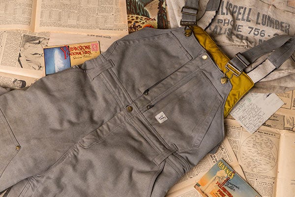 Gray work overalls with a front zipper and yellow lining laid on a background of scattered vintage book pages and magazines.