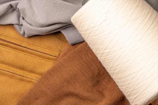 Close-up of layered Smith + Rogue fabrics featuring gray Pima cotton t-shirt material, tan Original Canvas fabric, brown Skoltec fabric, and spools of white Pima cotton thread.