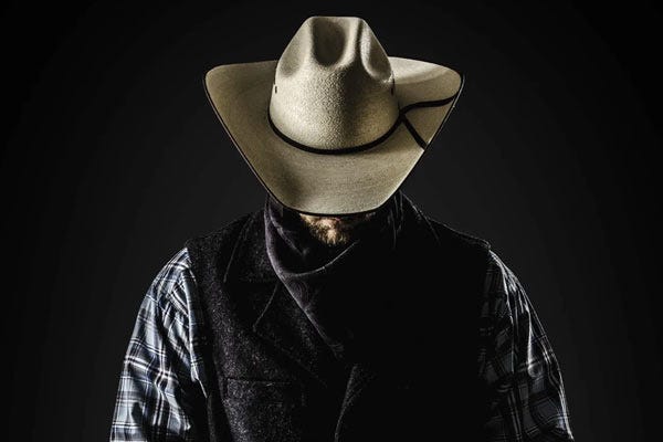 A man wearing a straw cowboy hat, plaid shirt, and dark vest stands against a black background with his head tilted downward.