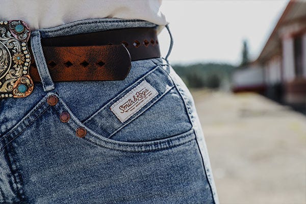 Close‑up of western jeans showing front pocket with stitched brand label, leather belt, and decorative belt buckle.