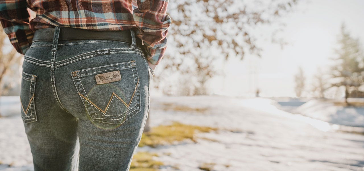 A woman stands facing away from the camera wearing wrangler jeans and a plaid shirts while snow covers part of the dormant grass in the winter