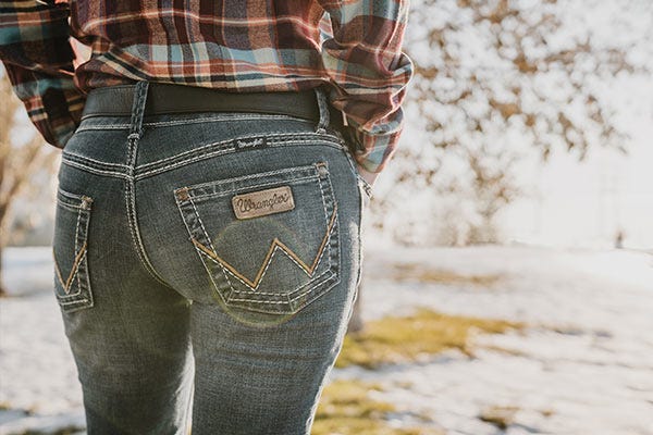 A woman stands facing away from the camera wearing wrangler jeans and a plaid shirts while snow covers part of the dormant grass in the winter