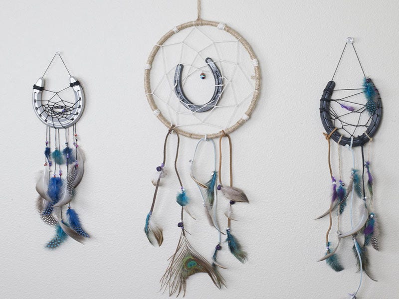Three handmade dreamcatchers hanging on a wall, each featuring a metal horseshoe woven into the center and decorated with string and hanging feathers.