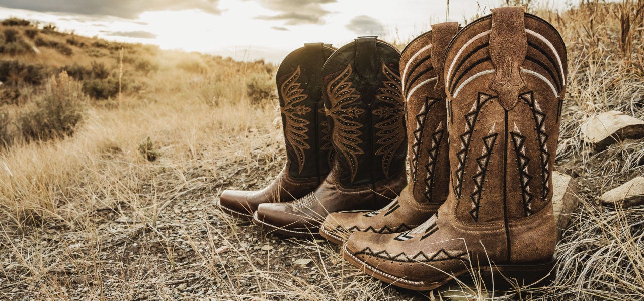 Two paris of empty cowboy boots sit side by side on a dirt and dry grass hill at sunset