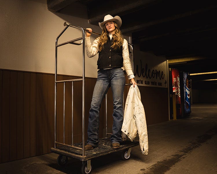 A woman wearing a cowboy hat, vest, jeans, and boots stands on a rolling luggage cart in a dimly lit hallway, holding a light‑colored jacket.