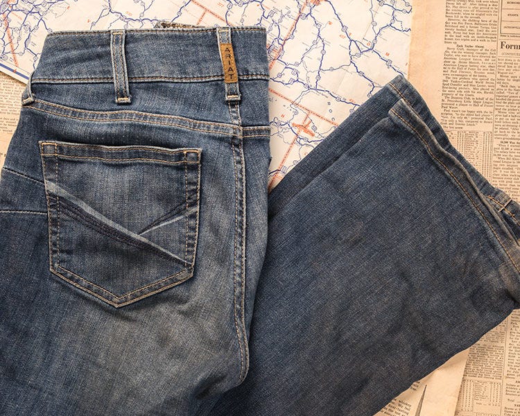 A pair of blue Ariat denim jeans laid flat, showing the back pocket and folded leg.