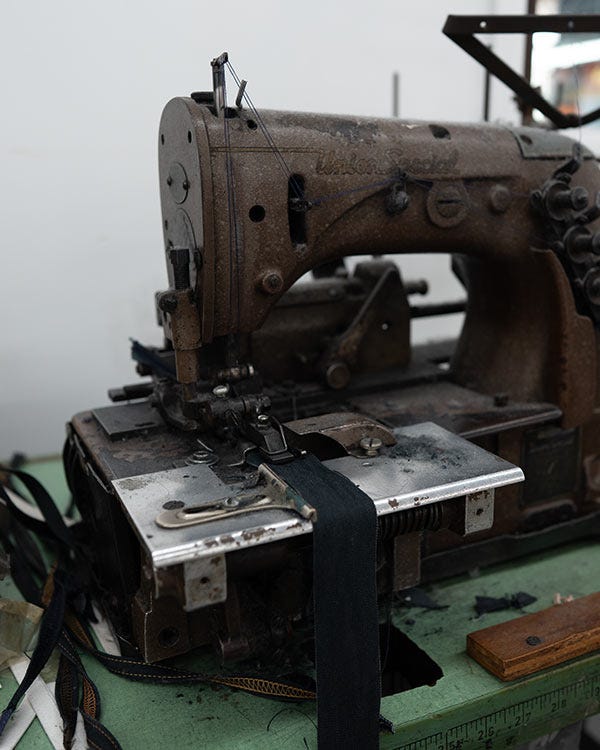 An industrial sewing machine stitches dark hemp fabric on a workshop table, with tools and fabric strips nearby