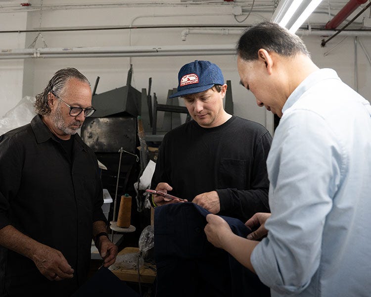 Three people stand in a workshop examining dark fabric materials together under fluorescent lighting, with industrial equipment and worktables visible in the background.