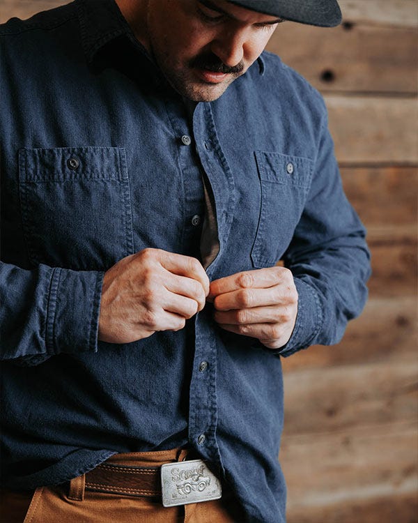 Person buttoning a dark blue hemp button-up shirt with chest pockets while standing indoors against a wooden wall.