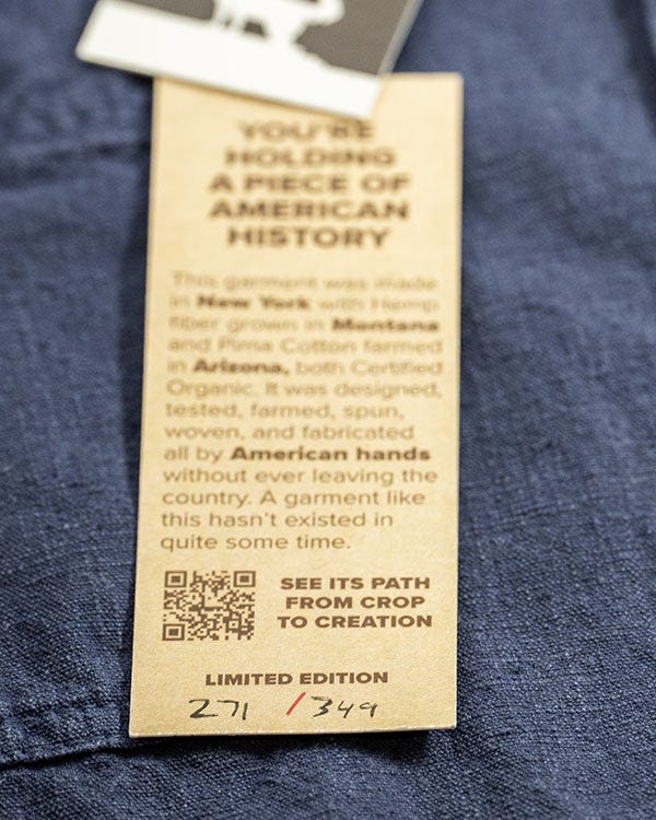 Close-up of a limited edition Benton shirt paper tag attached to dark blue hemp fabric, featuring product information, a QR code, and handwritten edition numbering