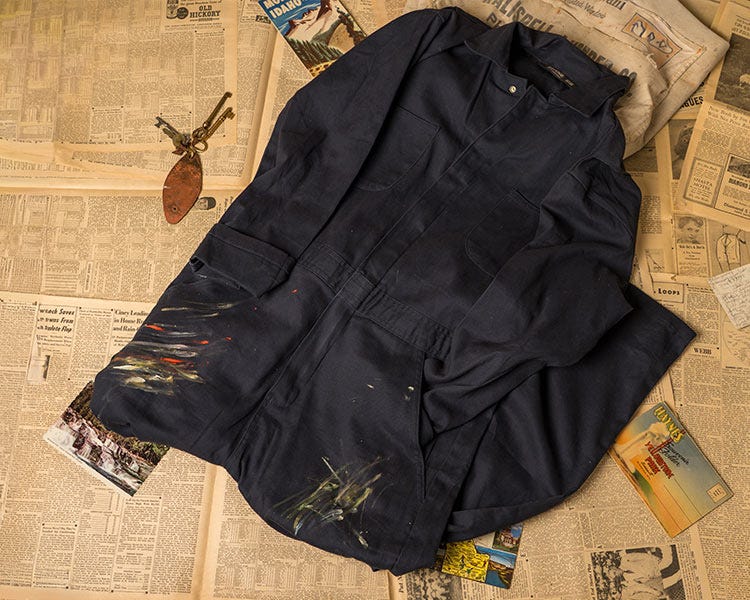 A pair of dark blue Berne Coveralls with paint smeared on the edges sitting on top of vintage newspapers, hotel keys, and postcards.