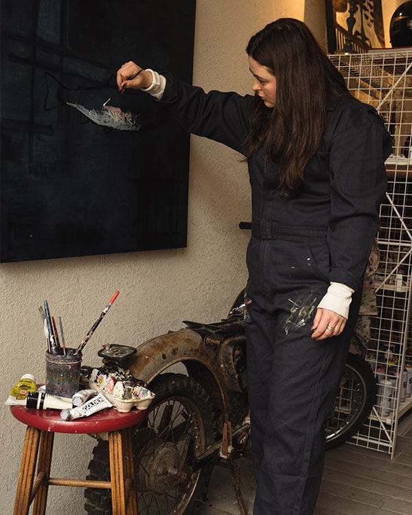 Maddie wearing Berne dark work coveralls painting on a canvas in a studio, with brushes, tools, and a vintage motorcycle nearby.