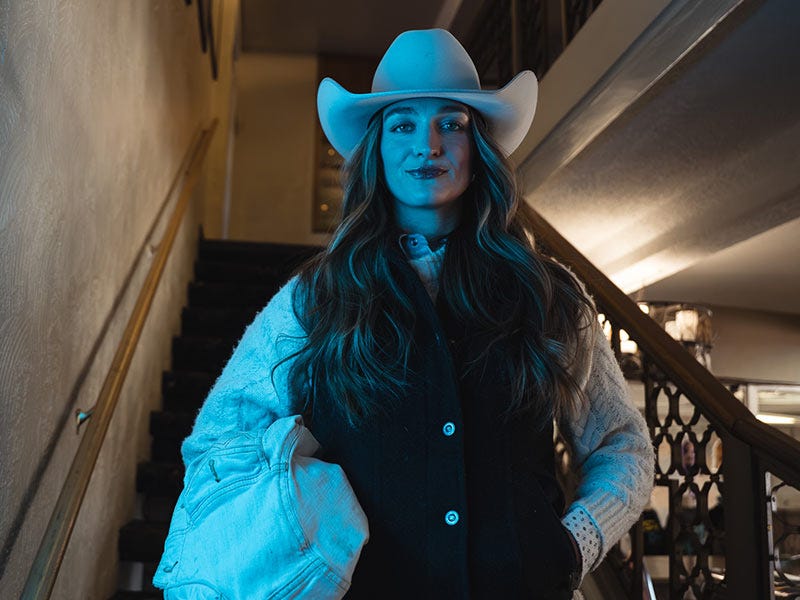 Caroline wearing a white Serratelli cowboy hat, dark vest, and light jacket standing indoors at the base of a staircase, lit with blue-toned lighting.