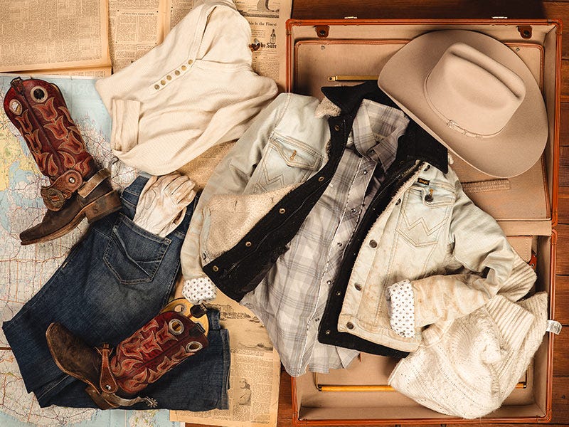 A full outfit spilling out of the suitcase across vintage newspaper. The outfit includes a cowboy hat, jacket, shirts, cowboy boots, ariat jeans, and gloves.