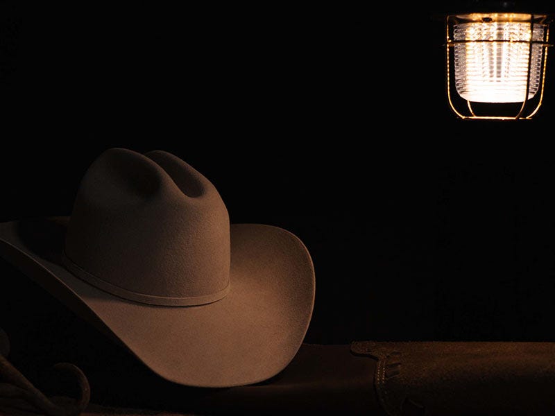 A brown cowboy hat resting on a wooden surface under a warm hanging lantern in a dark setting.