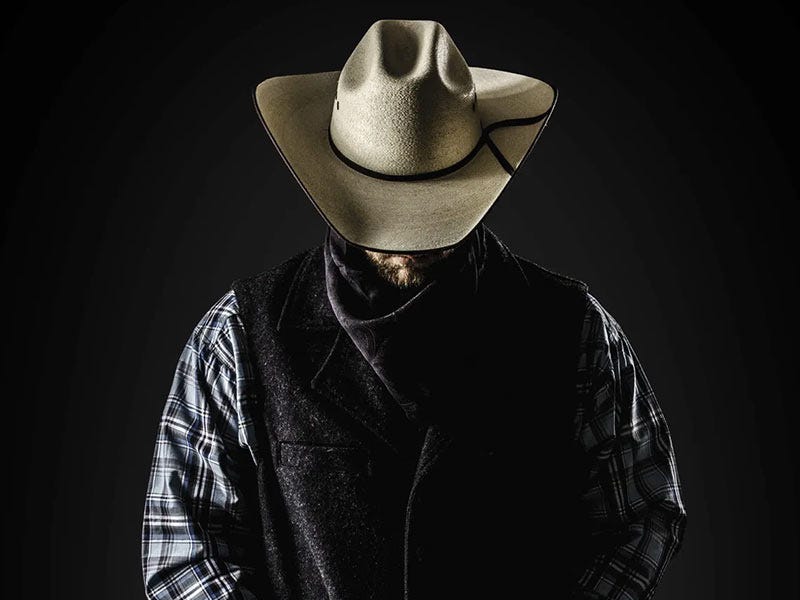 Man wearing a cowboy hat, plaid shirt, and dark vest, standing against a dark background with his face in shadow.