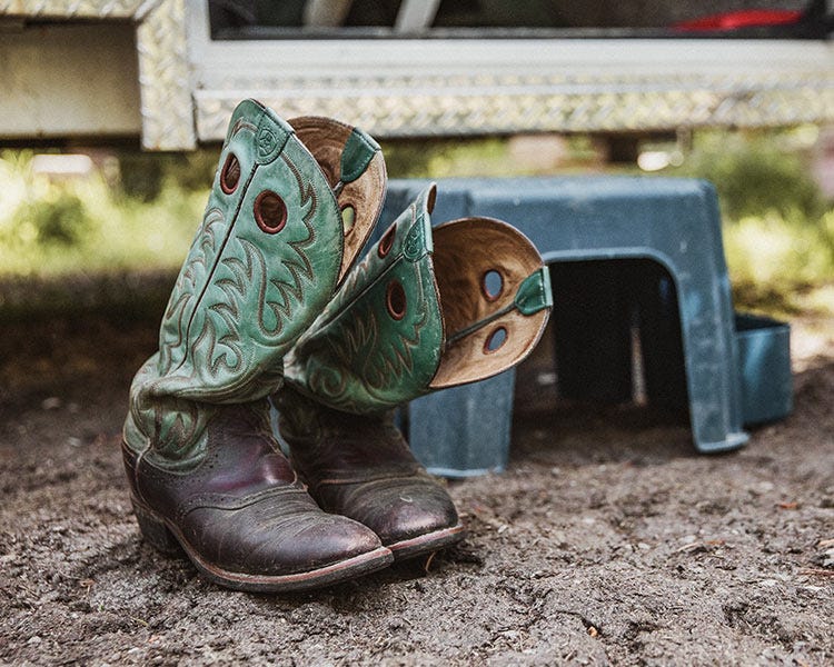 A pair of old teal cowboy boots sitting in front of a blue stool on the dirt. They are slumped with age.