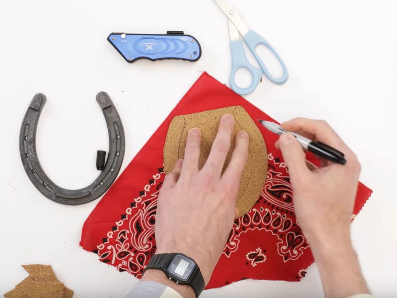 Hands tracing a cork horseshoe shape onto a red bandana with a marker, with scissors, a utility knife, and a metal horseshoe nearby.