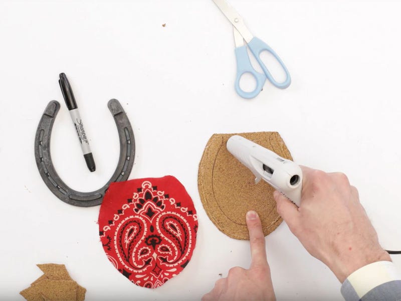 Person applying hot glue to a cork horseshoe shape, with a cut bandana piece, scissors, marker, and metal horseshoe on a white work surface.