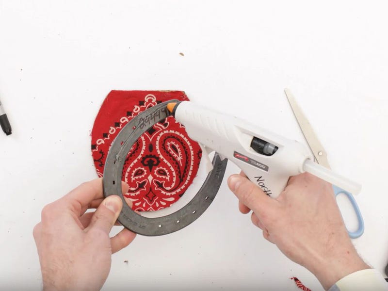 Person applying hot glue to a metal horseshoe attached to a bandana‑covered cork base on a white work surface.