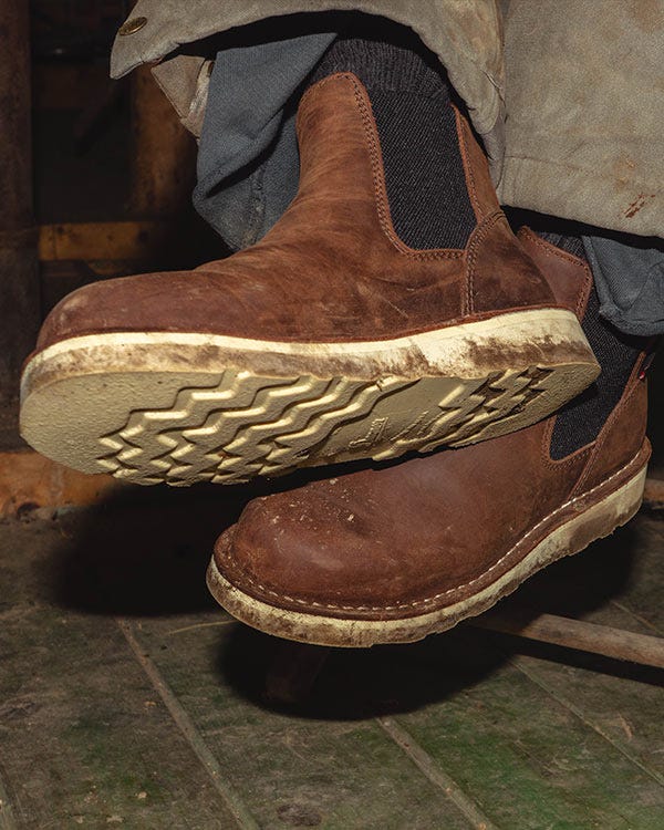 Close-up of brown Danner Bull Run Chelsea boots with elastic side panels and worn soles, shown against a rustic indoor floor with pant hems visible.