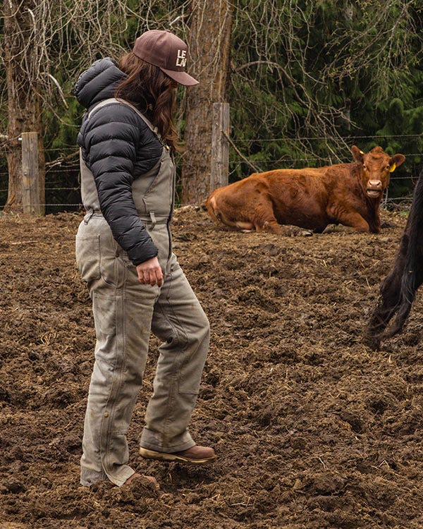 Maddie wearing a Smith + Rogue 777 puffy jacket, Smith + Rogue bib overalls, and brown Danner Chelsea boots standing in a muddy pasture near a cow and wooden fence posts.