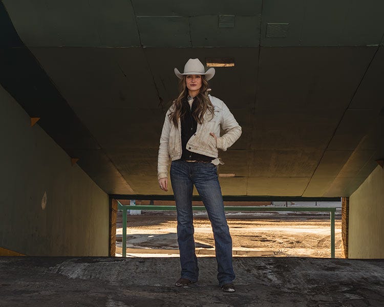 Woman wearing a light-colored cowboy hat, jacket, and jeans standing in a concrete underpass, facing the camera with one hand in their pocket