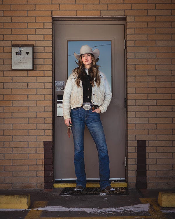 Woman wearing a light-colored cowboy hat, jacket, and jeans standing in a doorway against a brick wall, with one hand in their pocket.