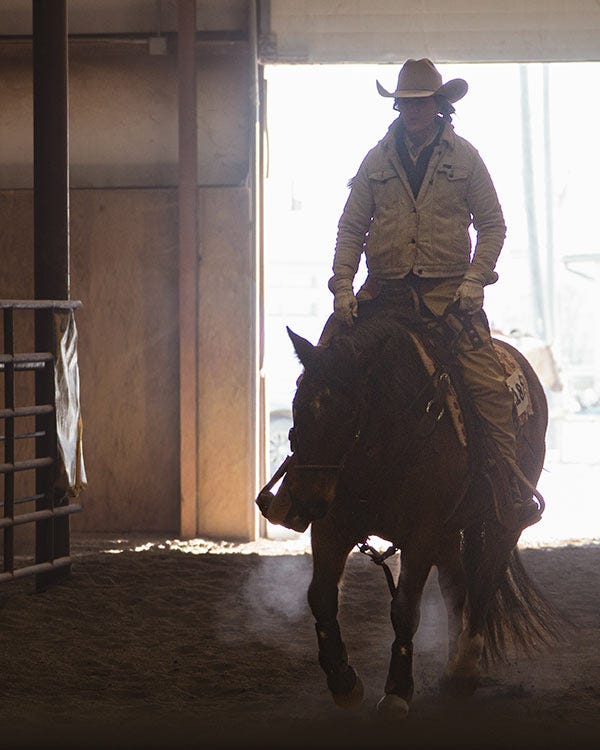 Woman wearing a cowboy hat and gloves riding a dark-colored horse inside a barn, backlit by daylight coming through an open doorway.