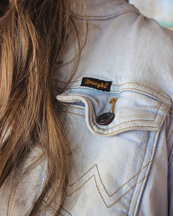 Close-up of the shoulder of a light-wash denim jacket with a buttoned flap pocket and a Wrangler brand label, with long hair visible along the shoulder.