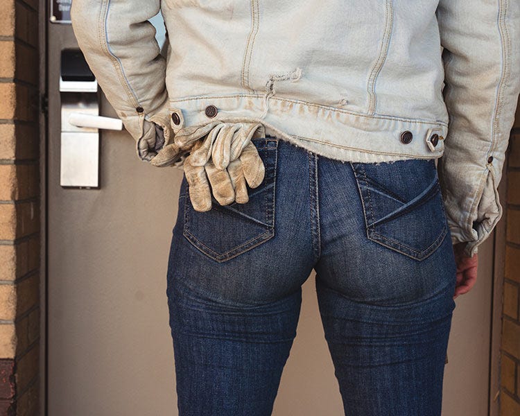 Rear view of a person wearing blue jeans and a light-colored jacket, with a pair of work gloves tucked into the back pocket, standing in a doorway.