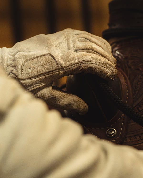 Close-up of a person wearing a light-colored leather work glove, resting their hand on a brown leather saddle in a warmly lit setting