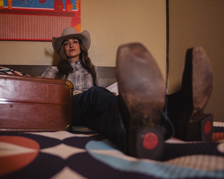 Woman wearing a cowboy hat and boots reclining on a bed with their feet toward the camera, with a suitcase beside them in a warmly lit room.