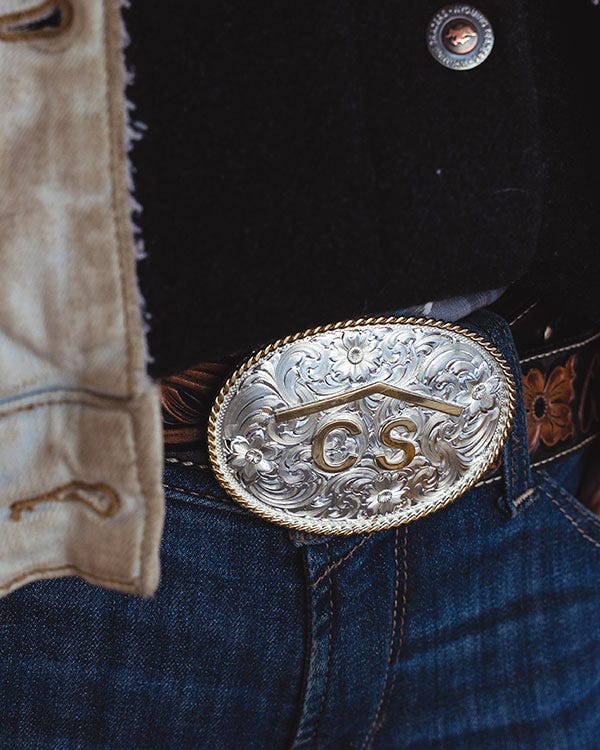 &ldquo;Close-up of an engraved silver belt buckle with the letters &lsquo;C S,&rsquo; worn with blue jeans, a leather belt, and a light-colored jacket.
