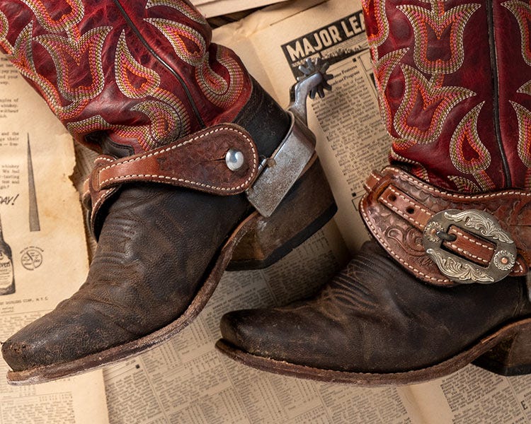 A pair of worn leather cowboy boots with red embroidered shafts and metal spurs, resting on a newspaper background.