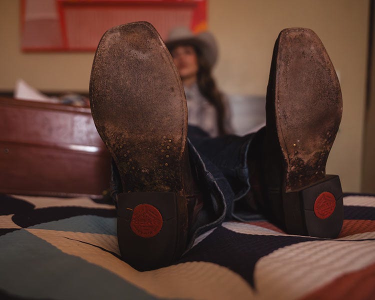 The worn soles of a pair of boots facing the camera, with a person seated out of focus in the background.