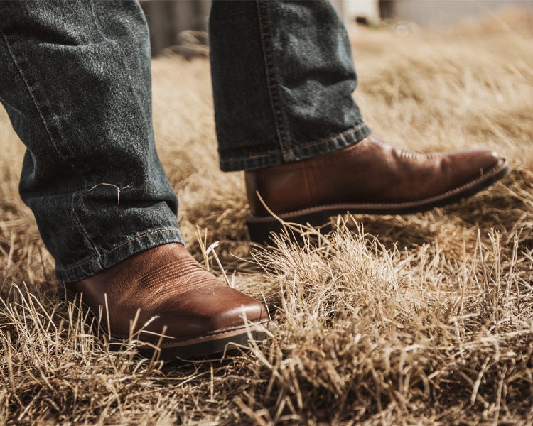 A close up of Justin boots and jeans in field with brown grass